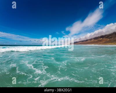Türkisfarbene Wellen schlagen am Sandstrand von Fuerteventura unter einem klaren blauen Himmel und schaffen eine ruhige Küstenatmosphäre. Stockfoto