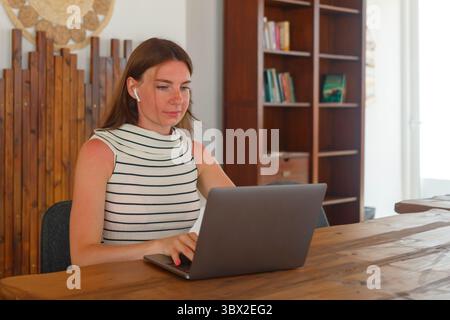 Die junge Frau konzentriert sich auf ihren Laptop in einem hellen und modernen Arbeitsbereich und hört Musik über kabellose Kopfhörer, während sie an einem Holztisch sitzt. Stockfoto