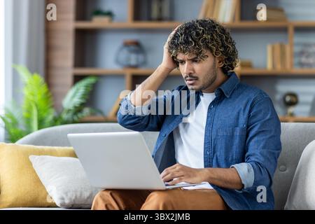 Ein besorgter Mann mit lockigen Haaren schaut auf einen Laptop und berührt seinen Kopf mit einem Ausdruck der Sorge. Stockfoto