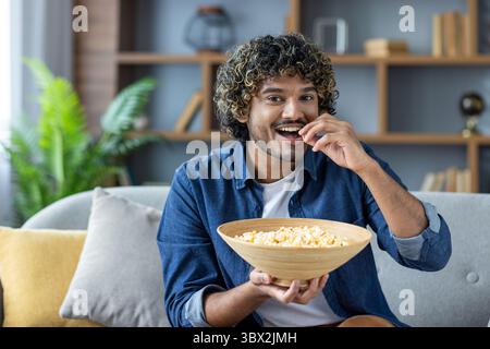 Ein lächelnder Mann genießt Popcorn, während er sich zu Hause auf einem Sofa entspannt. Er ist glücklich und isst einen Snack. Stockfoto