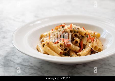 Köstliche italienische Rigatoni-Pasta mit farbenfrohem Gemüse, Karotten und geriebenem Parmesankäse, serviert in einer weißen Schüssel auf Marmoroberfläche. Stockfoto