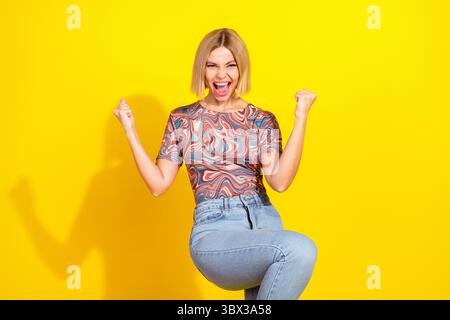 Die aufgeregte junge blonde Frau, die Fäuste zum Feiern hebt, trägt ein farbenfrohes lässiges Outfit vor leuchtendem gelbem Hintergrund Stockfoto