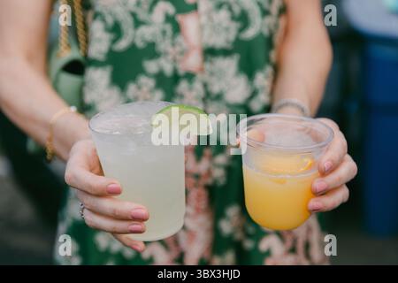 Zwei bunte Getränke werden von einer Person gehalten, umgeben von einer lebhaften Atmosphäre im Freien bei einer Sommerveranstaltung. Stockfoto