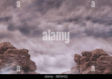 Flaches Design mit zwei rotbraunen Sandsteinformationen, die unter grauen Sturmwolken aufsteigen. Wüstenlandschaft, geologische Formation, Wettergestein, Ruggedterr Stockfoto