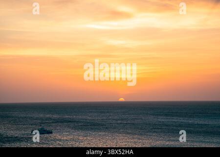 Goldene Farbtöne erfüllen den Himmel, während die Sonne über dem friedlichen Ozean untergeht und ein kleines Boot leise dahintreibt. Stockfoto
