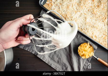 Hand hält einen Ständer Mixer Schneebesen-Aufsatz bedeckt mit verschlagenen Eiweiß: Geschlagenes Eiweiß auf einem Schneebesen mit kandierter Ananas und geröstetem Raspeln Stockfoto