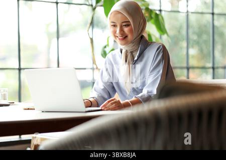 Junge asiatische indonesische Geschäftsfrau, die Hijab trägt und mit Laptop arbeitet, in einem Café neben dem Fenster sitzt Stockfoto