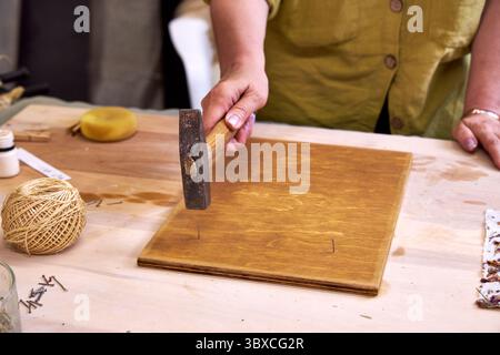 Nahaufnahme einer Frau, die Nägel in ein gebeiztes Holzbrett hämmert, für handgefertigte Dekorationen. Teil eines umweltfreundlichen DIY-Wandpaneelprojekts mit Naturpapier und Ma Stockfoto