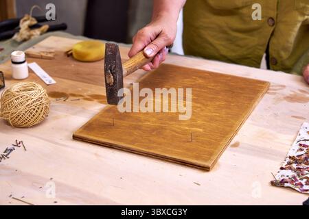 Nahaufnahme einer Frau, die Nägel in ein gebeiztes Holzbrett hämmert, für handgefertigte Dekorationen. Teil eines umweltfreundlichen DIY-Wandpaneelprojekts mit Naturpapier und Ma Stockfoto