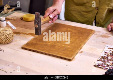 Nahaufnahme einer Frau, die Nägel in ein gebeiztes Holzbrett hämmert, für handgefertigte Dekorationen. Teil eines umweltfreundlichen DIY-Wandpaneelprojekts mit Naturpapier und Ma Stockfoto