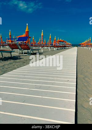 Ein weißer Holzweg führt zum Meer entlang von Liegestühlen und Sonnenschirmen am Sandstrand. Leere Liegestühle am Sandstrand. Stockfoto