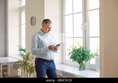 Ein konzentrierter, reifer Mann zu Hause, der auf den Bildschirm des Telefons blickt, im Stehen steht und Apps für mobile Geräte durchsucht Stockfoto