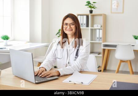 Glückliche lächelnde junge weibliche Hausarzt-Klinik-Ärztin am Laptop, Videoanruf, elektronische medizinische Aufzeichnungen Stockfoto