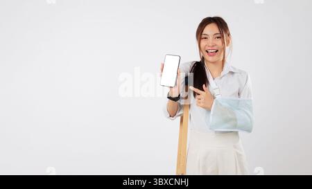 Verletzte asiatische Frau mit gebrochenem Arm, in einem Gips, das Telefon hält, benutzt mobile Unterstützung Stockfoto