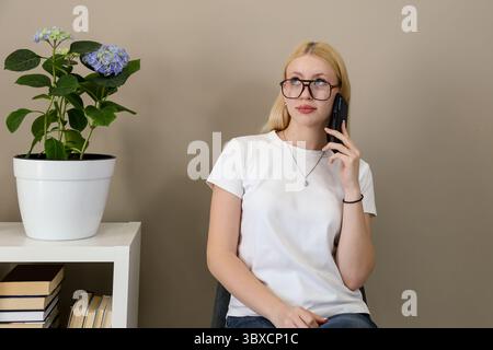 Die blonde Frau in einem weißen T-Shirt und einer Brille sitzt in einem gemütlichen Innenraum, hält ein Smartphone an ihrem Ohr, während sie nachdenklich aufblickt, neben einer Blüte Stockfoto