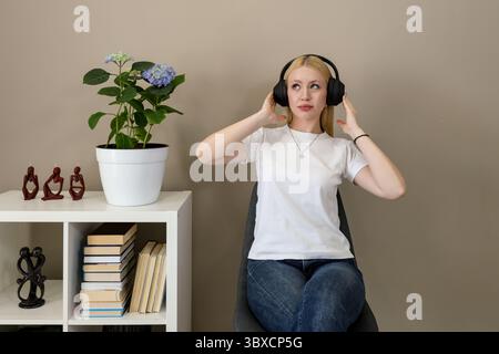 Die junge blonde Frau in weißem T-Shirt und Jeans sitzt bequem auf einem Stuhl, trägt große schwarze Kopfhörer und genießt Musik in einem gemütlichen, modernen Inter Stockfoto