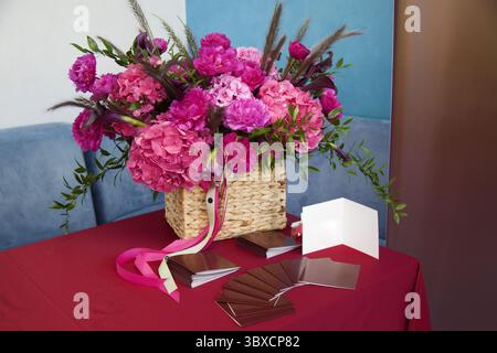 Üppiger Blumenstrauß aus rosafarbenen Pfingstrosen und Hortensien mit Grün in einem gewebten Korb auf einer roten Tischdecke, umgeben von leeren Grußkarten und Satinbändern, Stockfoto