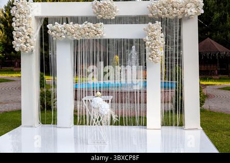 Hochzeiten im Freien mit einem modernen weißen Bogen, der mit weißen Rosen und Bändern verziert ist, vor einem Brunnen in einem malerischen garten Stockfoto