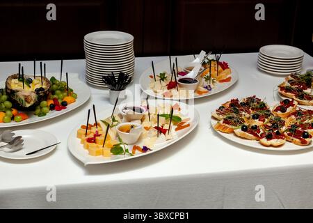 Gourmet-Catering-Einrichtung mit Käsewürfeln, Obstspießen und Bruschetta mit Oliven und Sprossen auf weißen Platten, bereit für Feiern oder sogar sogar Stockfoto