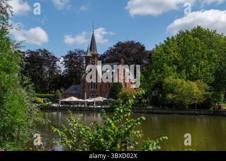 Diese bezaubernde Flussszene in Brügge verfügt über ein historisches Gebäude aus roten Backsteinen mit einem Turm, Caféschirmen am Wasser und üppiges Grün, das zum Entspannen einlädt Stockfoto