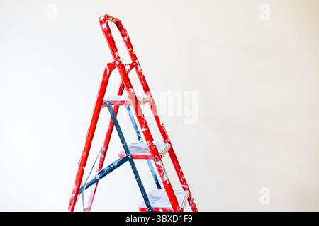 Eine rote Stufenleiter, die an einer sauberen weißen Wand anliegt, ist einsatzbereit. Eine rote Stufenleiter steht vor einer einfachen weißen Wand, perfekt für die Heimpflege Stockfoto