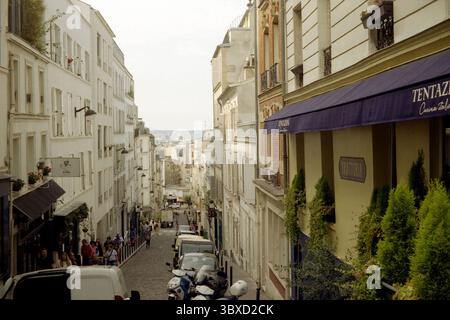 Offener Blick auf die Straße im Sommer auf die Rue Lepic in Montmartre, Paris, mit Fußgängern, geparkten Autos und Restaurant-Markise. aufgenommen auf 35-mm-Film. Stockfoto
