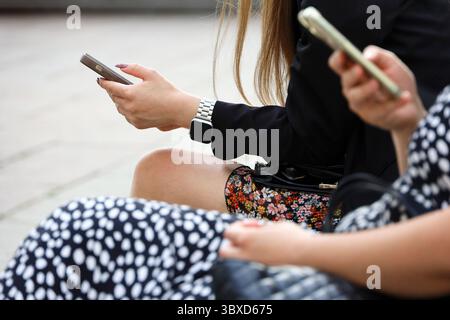 Menschen, die Smartphones auf der Straße in der Stadt nutzen. Mädchen sitzen im Sommer mit Handys in der Hand auf einer Bank Stockfoto