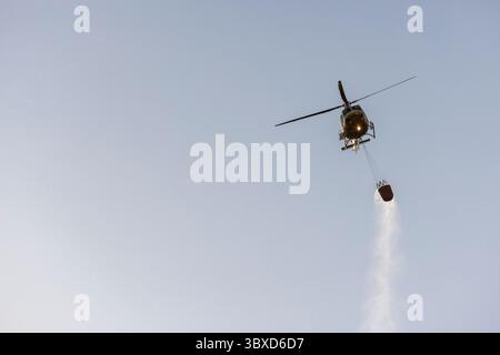 Allariz, Spanien, 16. Juli 2025: Ein gelb-schwarzer Feuerwehrhubschrauber fliegt am Himmel. Der Hubschrauber trägt einen Feuerwehrschlauch und fällt Stockfoto