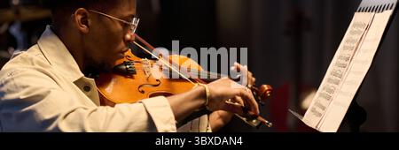 Junger erwachsener Schwarzer Mann, der Geige spielt, während er Noten am Stand liest, drinnen mit fokussiertem Ausdruck sitzt, eine Brille trägt und musikalische Fähigkeiten demonstriert Stockfoto