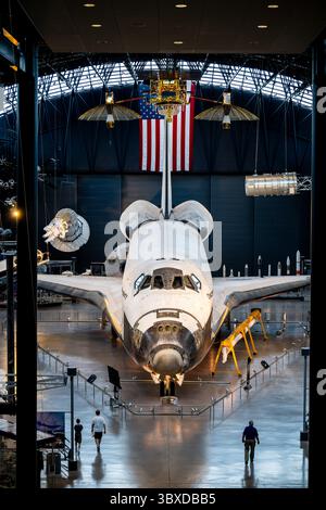 Space Shuttle Discovery Display im Steven F Udvar-Hazy Center in Chantilly Virginia | CHANTILLY, Virginia – Besucher spazieren unter der Nase des Space Shuttle Discovery im James S. McDonnell Space Hangar im Steven F. Udvar-Hazy Center, das von der 78 Fuß (24 Meter) langen Flügelspanne des Orbiters im Schatten steht. Die Discovery flog zwischen 1984 und 2011 39 Missionen, das meiste aller Space Shuttles, und dient heute als erhaltenes „Fahrzeug der Aufzeichnung“ der NASA. Stockfoto