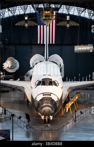 Space Shuttle Discovery Display im Steven F Udvar-Hazy Center in Chantilly Virginia | CHANTILLY, Virginia – zwei Besucher stehen unter der Nase des Space Shuttle Discovery, während Satelliten-Hardware und eine amerikanische Flagge über dem James S. McDonnell Space Hangar hängen. Die Entdeckung, mit 39 Missionen der meistgeflogene Orbiter in der Geschichte der NASA, ist im Steven F. Udvar-Hazy Center als „Fahrzeug der Aufzeichnung“ des Programms erhalten. Stockfoto
