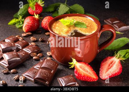 Genießen Sie dieses gemütliche Bild mit einer warmen Tasse Kaffee vor hölzernem Hintergrund, begleitet von frischen Erdbeeren und einem dekadenten Schokoladenstück Stockfoto