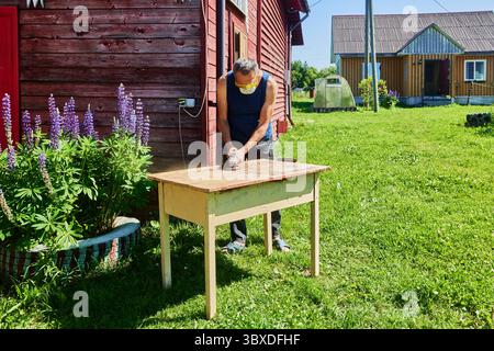 Holztisch mit Winkelschleifer im Freien schleifen, Hobbyarbeit im Hof in der Nähe von Holzhäusern an sonnigen Sommertagen auf dem Land. Stockfoto