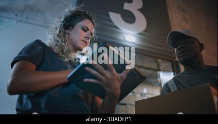 Weibliche Manager scannt Pakete mit Barcode-Scanner, mit Digital Tablet, arbeitet nachts. Verschiedene Mitarbeiter, die Kartonschachteln aus dem Logistiklager transportieren. Online-Bestellungen, Lieferung. Zeitlupe. Stockfoto