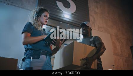Verschiedene Mitarbeiter, die Kartonschachteln aus dem Logistics Warehouse transportieren und nachts arbeiten. Weibliche Manager scannt Pakete mit Barcode-Scanner mit Digital Tablet. Online-Bestellungen, Lieferung. Zeitlupe. Stockfoto