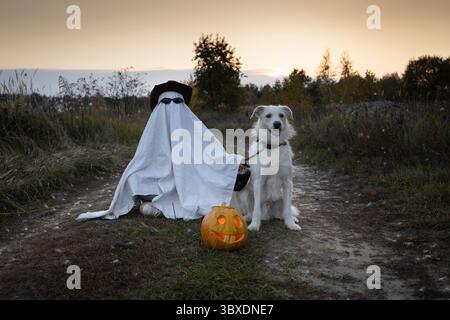 Eine nicht erkennbare Person in weißer Decke, Hut und schwarzer Brille geht mit einem weißen Hund durch den Park. Ein Teenager im Geisterkostüm geht mit seinem Haustier auf Hallo Stockfoto