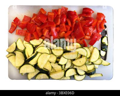 In Halbmonde geschnittene Zucchini und gehackte rote Paprika ruhen auf einem weißen Schneidebrett. Stockfoto