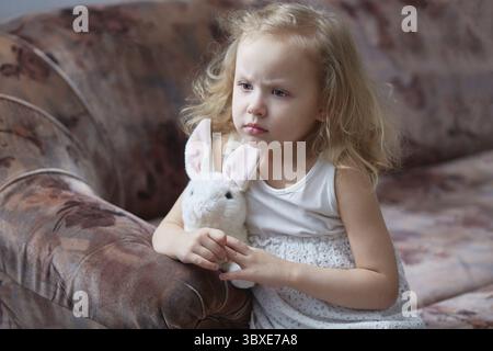 Das lockige Mädchen ist traurig, wenn es zu Hause auf der Couch sitzt und ein weiches Spielzeug umarmt Stockfoto