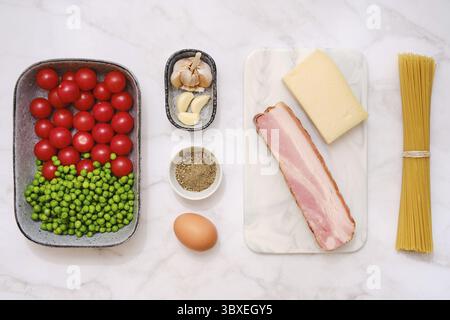 Blick von oben auf frische Kirschtomaten, grüne Erbsen, Räucherspeck, Knoblauch, Parmesankäse, Spaghetti und ein Ei auf einer hellen Küchentheke Stockfoto