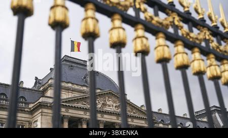Der Königspalast in Brüssel, Belgien. Blick durch den Metallzaun mit goldenen Details. Nationalflagge des Königreichs Belgien winkt oben Stockfoto