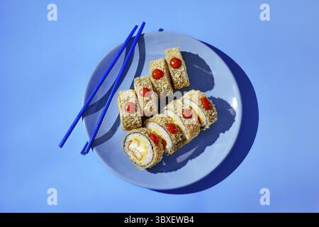 Frisch zubereitete Tempura-Maki-Sushi-Brötchen mit einer scharfen Sauce werden auf einem blauen Teller mit blauen Essstäbchen serviert, was eine lebendige kulinarische Szene schafft Stockfoto