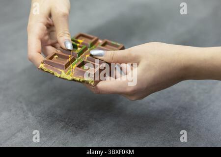 Weibliche Hände brechen ein Stück Dubai Schokolade auf grauem Hintergrund ab. Grüner Kadayif und Pistazien mit Pistazien im Trenddessert. Nahaufnahme, s Stockfoto