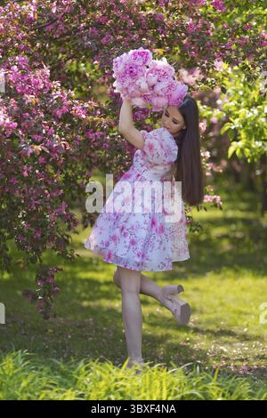 Glückliches, schönes Mädchen mit langen Haaren, Brünette, in einem hellrosa Kleid, mit einem Strauß großer rosafarbener Pfingstrosen, in der Garde neben rosa Blüten Stockfoto