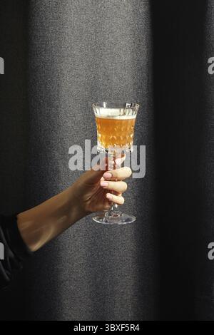 Weibliche Hand elegant hält Kristallglas gefüllt mit Whiskey sauer, Cocktail in bernsteinfarbenem Farbton im Kontrast mit dunklem strukturiertem Stoffhintergrund Stockfoto