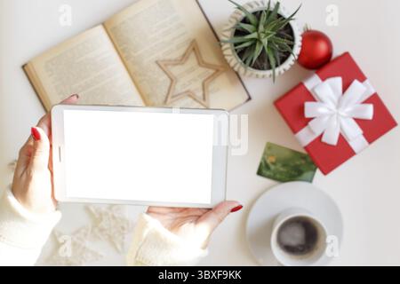 Schöne Frauenhände halten ein digitales Tablet über dem Tisch mit Kreditkarte, offenem Buch, roter Geschenkbox, Tasse Kaffee und saftiger Pflanze, weihnachtsdekor und Stockfoto