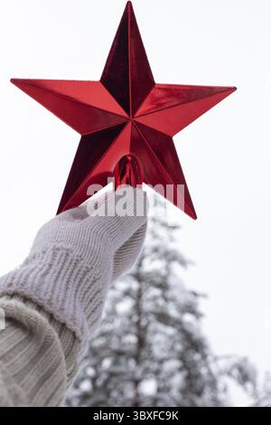Eine Hand in einem weißen gestrickten Fäustling hält einen roten Stern über einer Schneepflanze im Wald. Vertikal Stockfoto