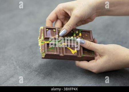 Weibliche Hände brechen ein Stück Dubai Schokolade auf grauem Hintergrund ab. Grüner Kadayif und Pistazien mit Pistazien im Trenddessert. Nahaufnahme, s Stockfoto