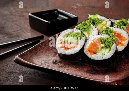 Appetitliche bunte Lachs- und Gurken-Maki-Sushi-Brötchen mit Tobiko und Salat, traditionell auf rustikalem Teller mit Sojasauce serviert. Japanische Küche Stockfoto