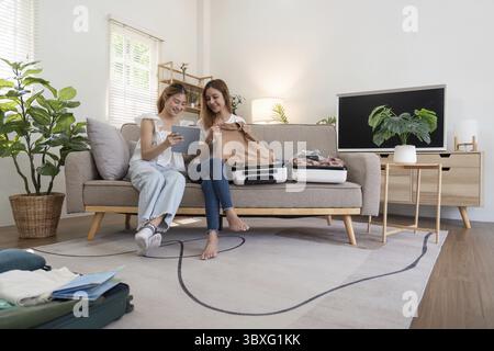 Eine junge asiatische Frau, die Kleider in den Koffer packt. Vorbereitung auf die Sommerferien. Zwei Frauen planen und mit dem Tablet für die Reisebuchung A Stockfoto
