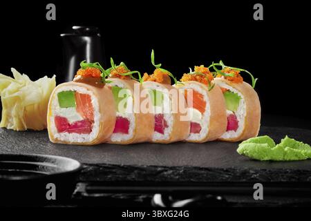 Gourmet-Sushi-Brötchen mit rohem Thunfisch, Lachs, Avocado und Frischkäse, verpackt in Mamenori mit scharfer Sauce und Tobikokaviar, serviert auf schwarzem Schieferbrett Stockfoto